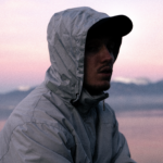 Portrait de Linf de profil, photo prise à l'extérieur à l'aube du jour qui se lève, il porte une veste à capuche et une casquette