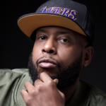 Portrait de Talib Kweli, le rappeur tient son menton et regarde droit dans l'objectif, il porte une casquette