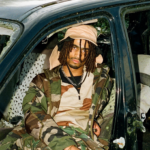 Portrait de l'artiste Toutencamo. Photo prise dans une voiture. L'artiste porte un couvre chef beige et des vêtements type treillis militaire.