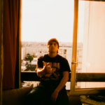 Le rappeur H JeuneCrack est pris en photo assis sur le rebord d'une fenêtre à l'intérieur d'un appartement, il point du doigt l'objectif de l'appareil photo